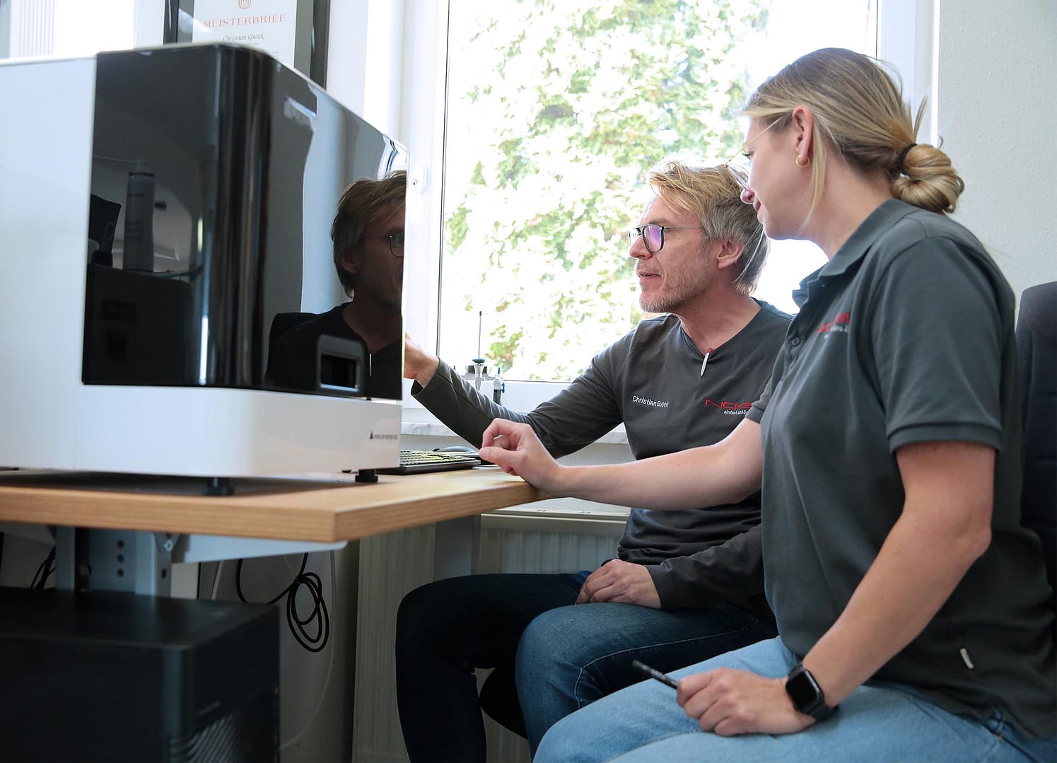 Zahntechniker und Zahntechnikerin sitzen vor einem Computer und einem 3D-Drucker für Zahnprothesen