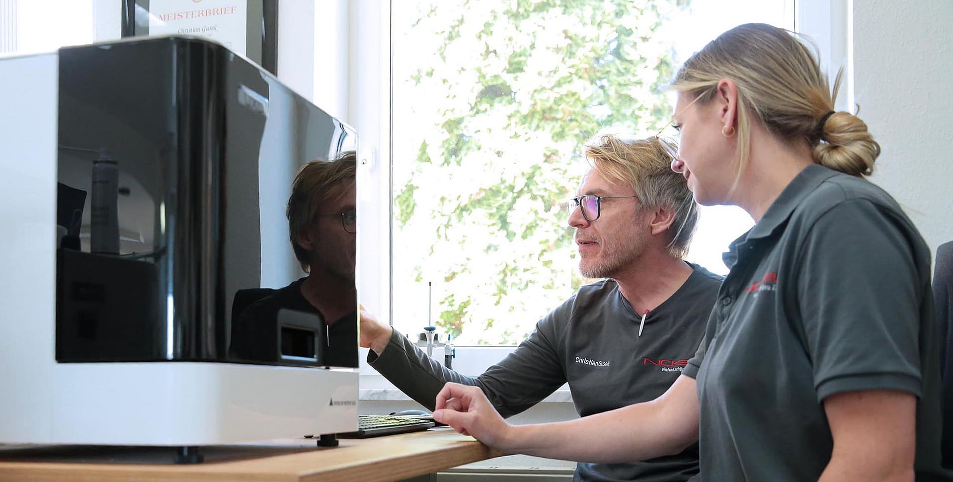 Zahntechniker und Zahntechnikerin sitzen vor einem Computer und einem 3D-Drucker für Zahnprothesen